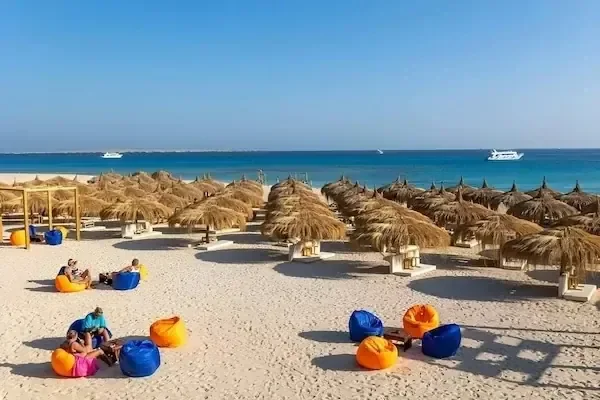 مظلات شاطئ من القش ومقاعد أكياس برتقالية وزرقاء موضوعة على رمال بيضاء تواجه البحر