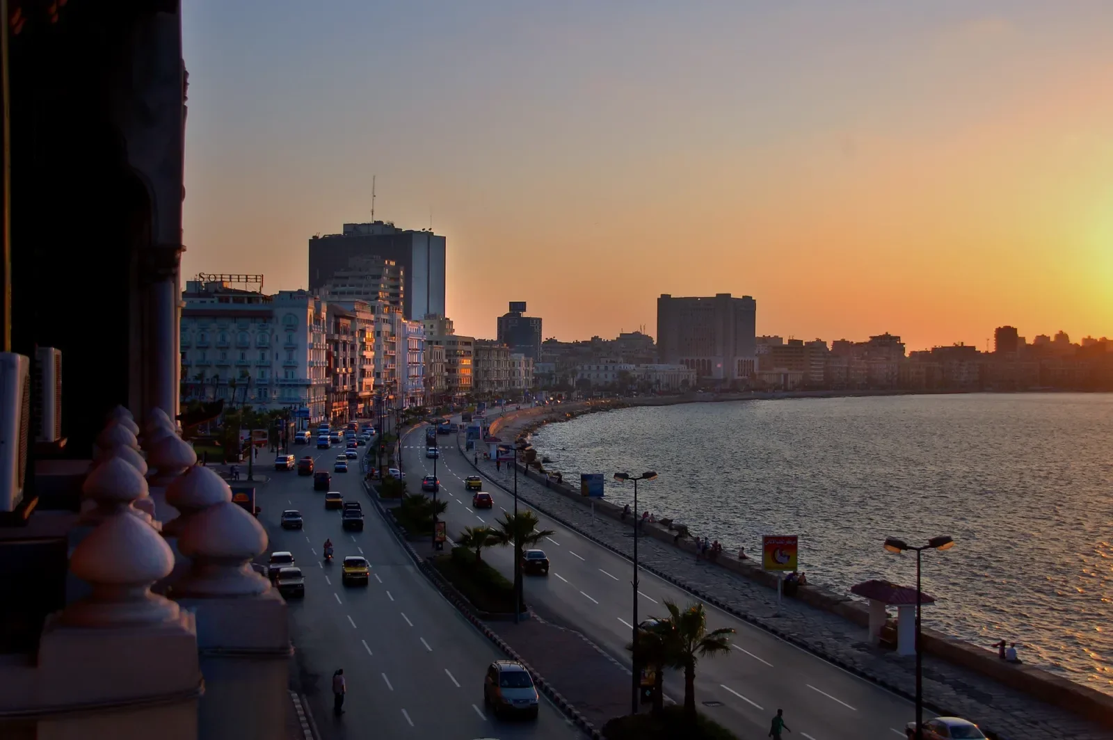A sunset view over a coastal boulevard with traffic and palm trees, seen from a balcony overlooking the sea.