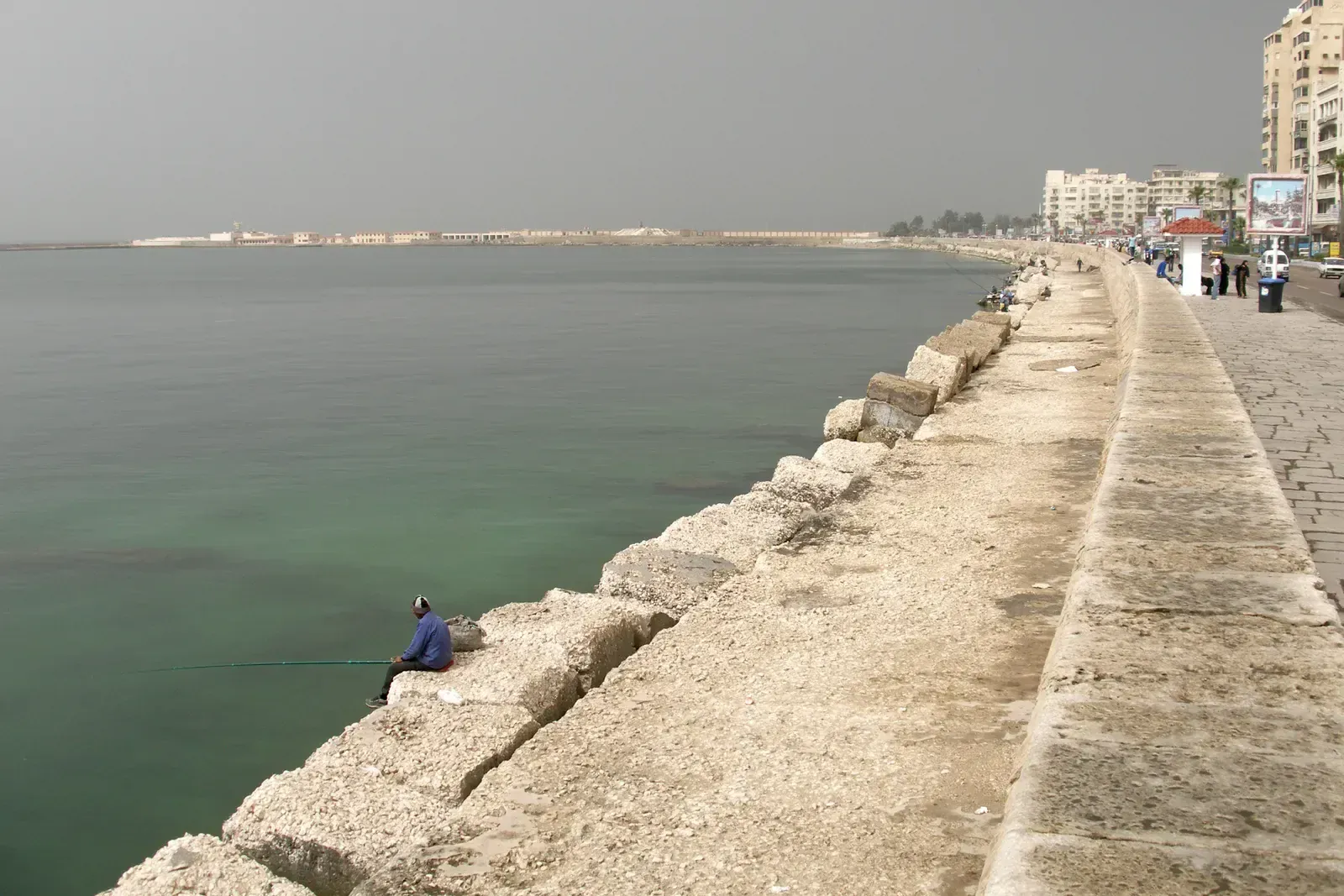 A man fishing with a rod from a stone seawall along a coastal promenade.