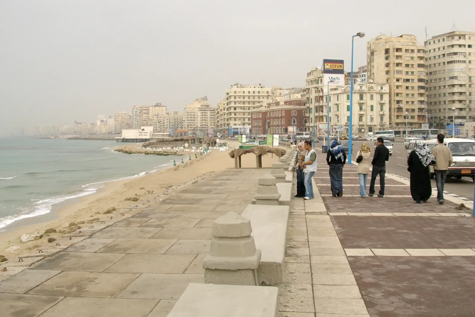 Pedestrians walking along a stone corniche seawall beside the sea with the city skyline behind them.