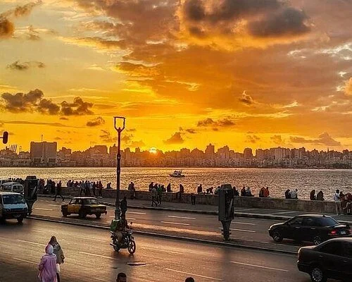 Silhouettes of pedestrians and traffic along the corniche at sunset, with the bay glowing orange behind them.