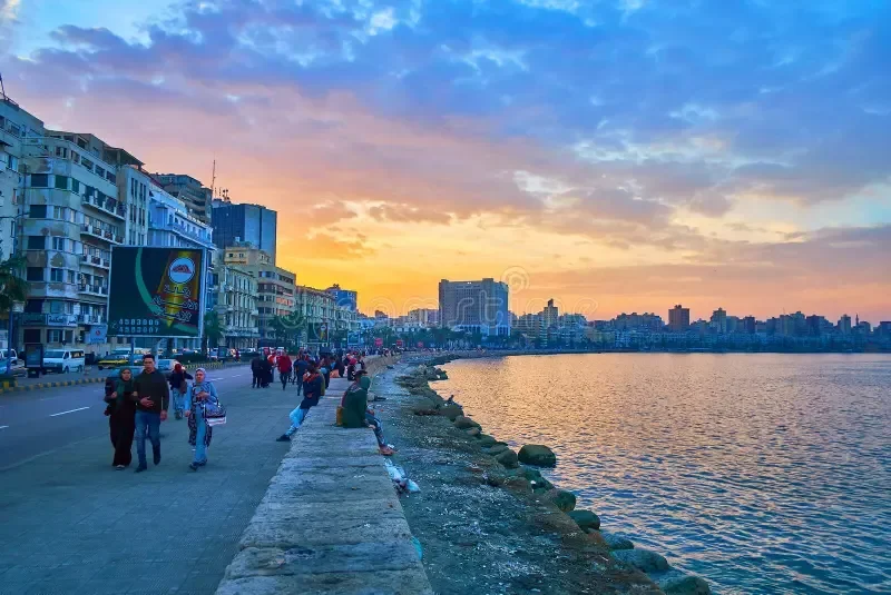 Pedestrians walking along the corniche seawall at sunset with the city skyline glowing behind them.