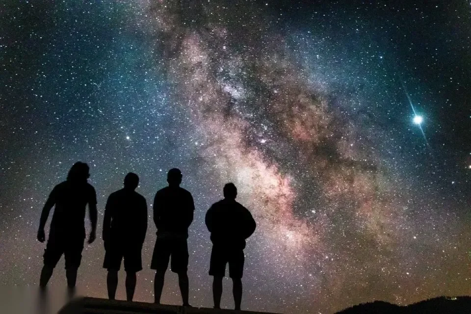 四个人的剪影站在山脊上，看银河横跨布满星星的沙漠夜空