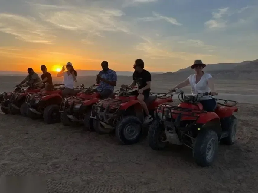 六名骑手驾驶红色全地形四轮摩托在沙漠中排成一列，夕阳落在远处山丘后