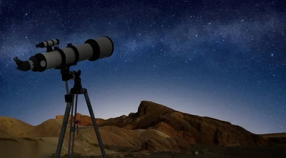 三脚架上的望远镜剪影映衬着布满星星的夜空，下方是岩石沙漠丘陵