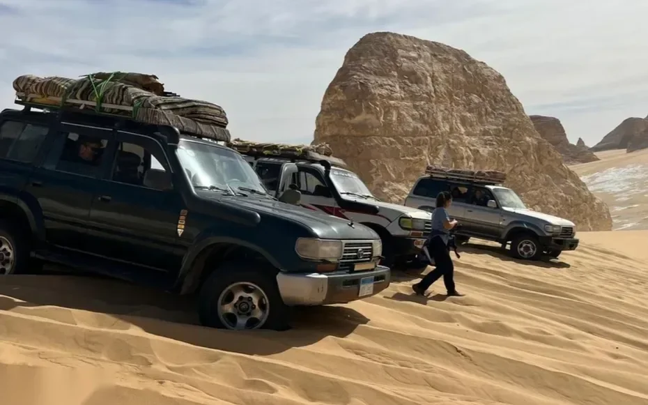 Drei Land Cruiser mit Matratzen beladen neben einem großen Felsblock, ein Guide geht vorbei