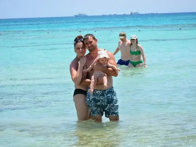 Gezin waadt in ondiep turquoise water, de vader houdt een baby vast terwijl een ander stel op de achtergrond staat