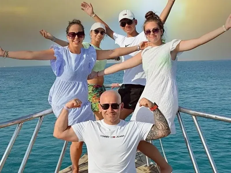 Gezin van vijf poseert met opgeheven armen op de boeg van een boot bij zonsondergang