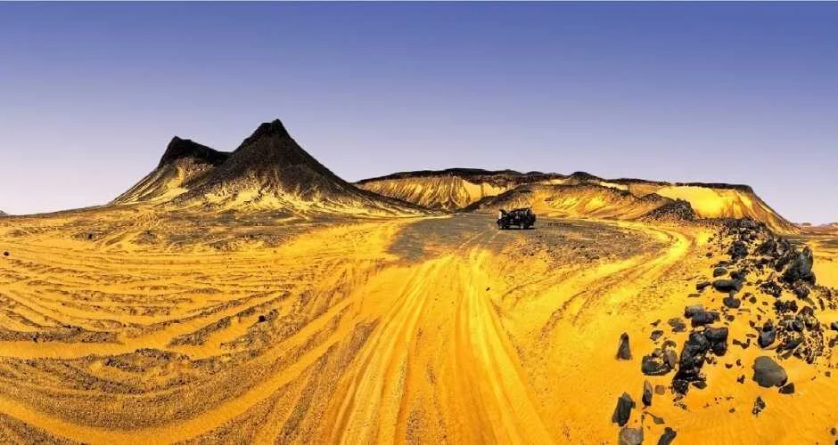 Vaste panorama désertique de pistes de sable doré menant vers des pics volcaniques noirs, un seul 4x4 visible au loin