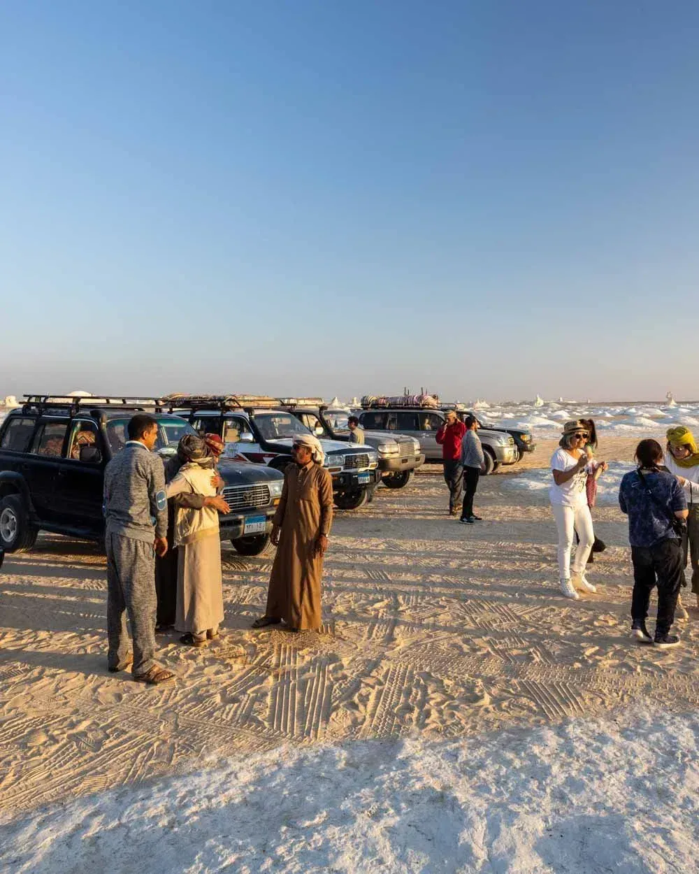 Touristes et guides bédouins près de 4x4 garés sur un sol blanc croûté de sel au coucher du soleil, désert Blanc
