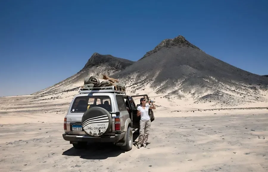 Femme prenant une photo avec un appareil, debout près d'un Land Cruiser 4x4 argenté garé devant des pics du désert noir