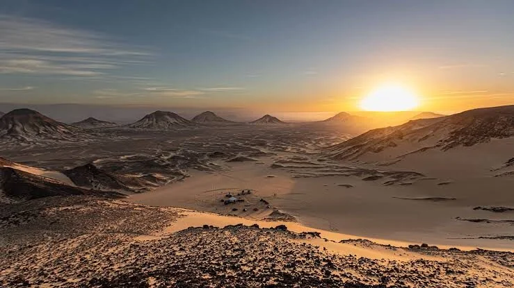 Vue aérienne d'un camp du désert avec plusieurs 4x4 garés au coucher du soleil, entouré des collines sombres du désert Noir