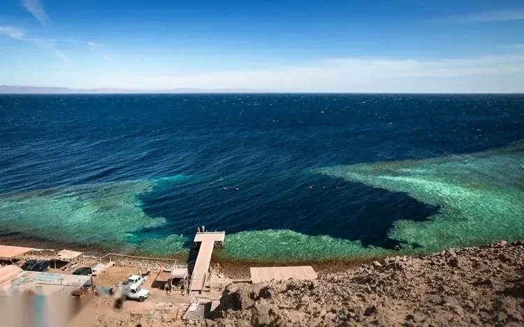 Légifelvétel a sziklaszirtről a dahabi Blue Hole zátonybemenetére, pickupokkal és kis partmenti épületekkel