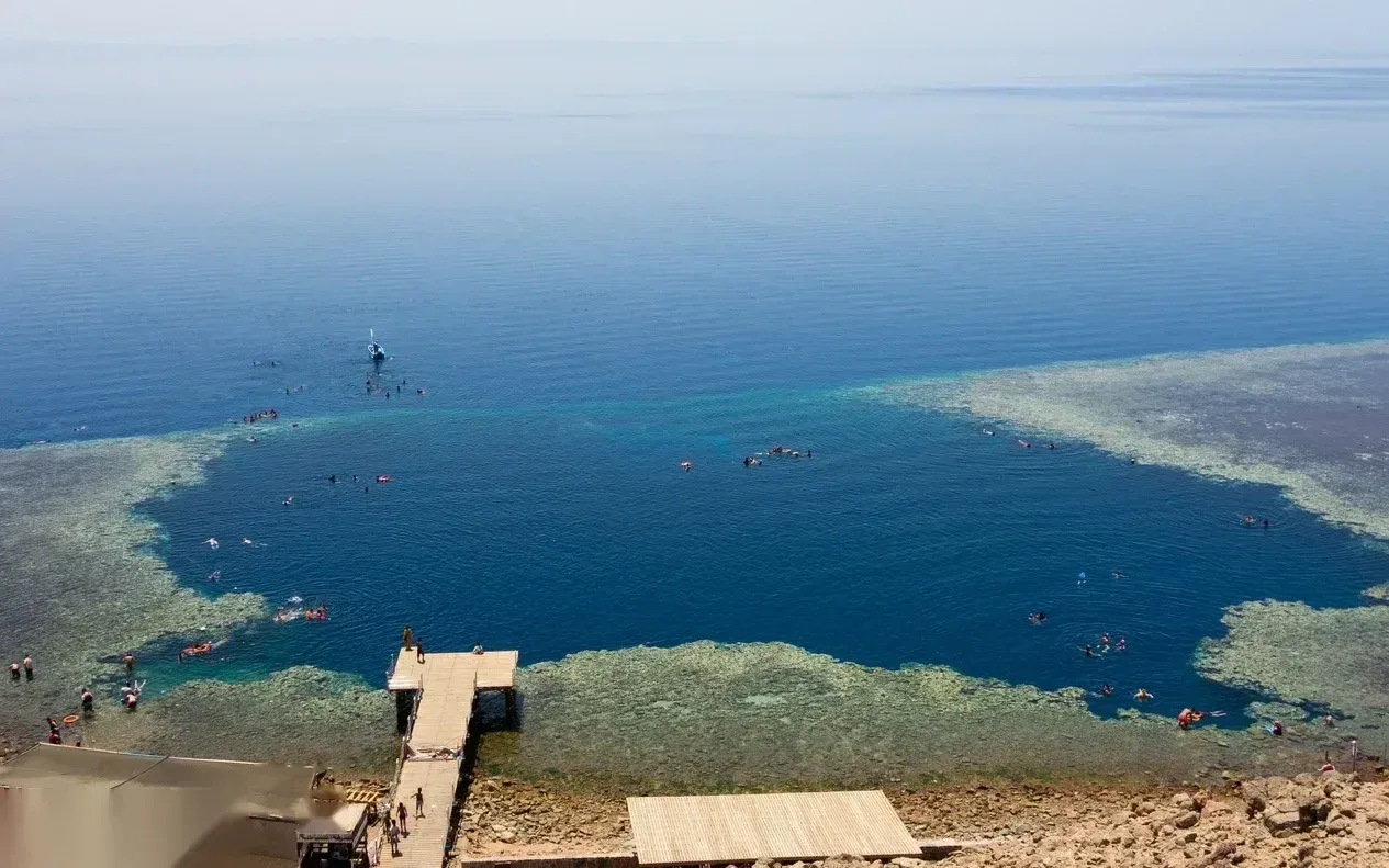 Tágabb magaslati nézet a Blue Hole lagúnájáról, amelyen több snorkelező szóródik szét a vízen és a zátonyon a móló közelében