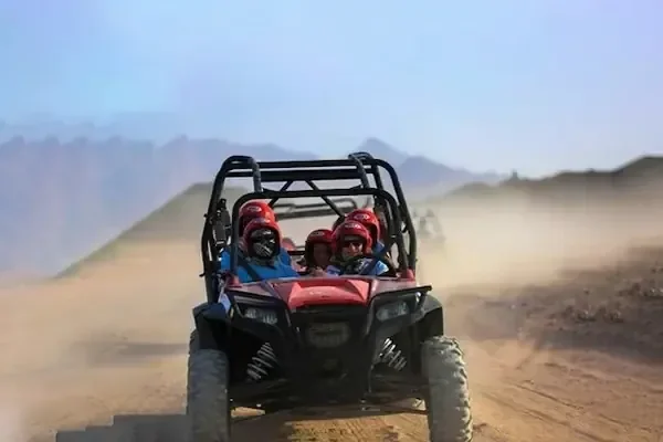 عربة كثبان حمراء وعليها راكبان بخوذتين تسير نحو الكاميرا على مسار صحراوي مترب، وخلفها جبال