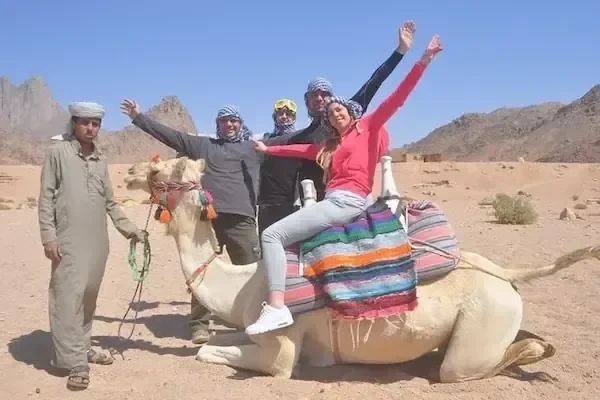 سائحان يركبان الجمال بقيادة مرشد بدوي عبر الصحراء وخلفهما جبال، وتظهر علامة مائية لموقع إلكتروني في أسفل الصورة