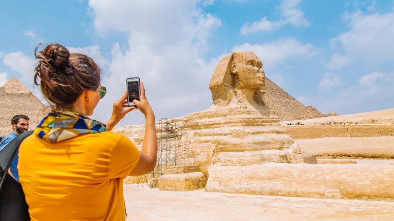 Vrouw fotografeert de Grote Sfinx met haar telefoon terwijl een man naast haar staat, piramides zichtbaar op de achtergrond