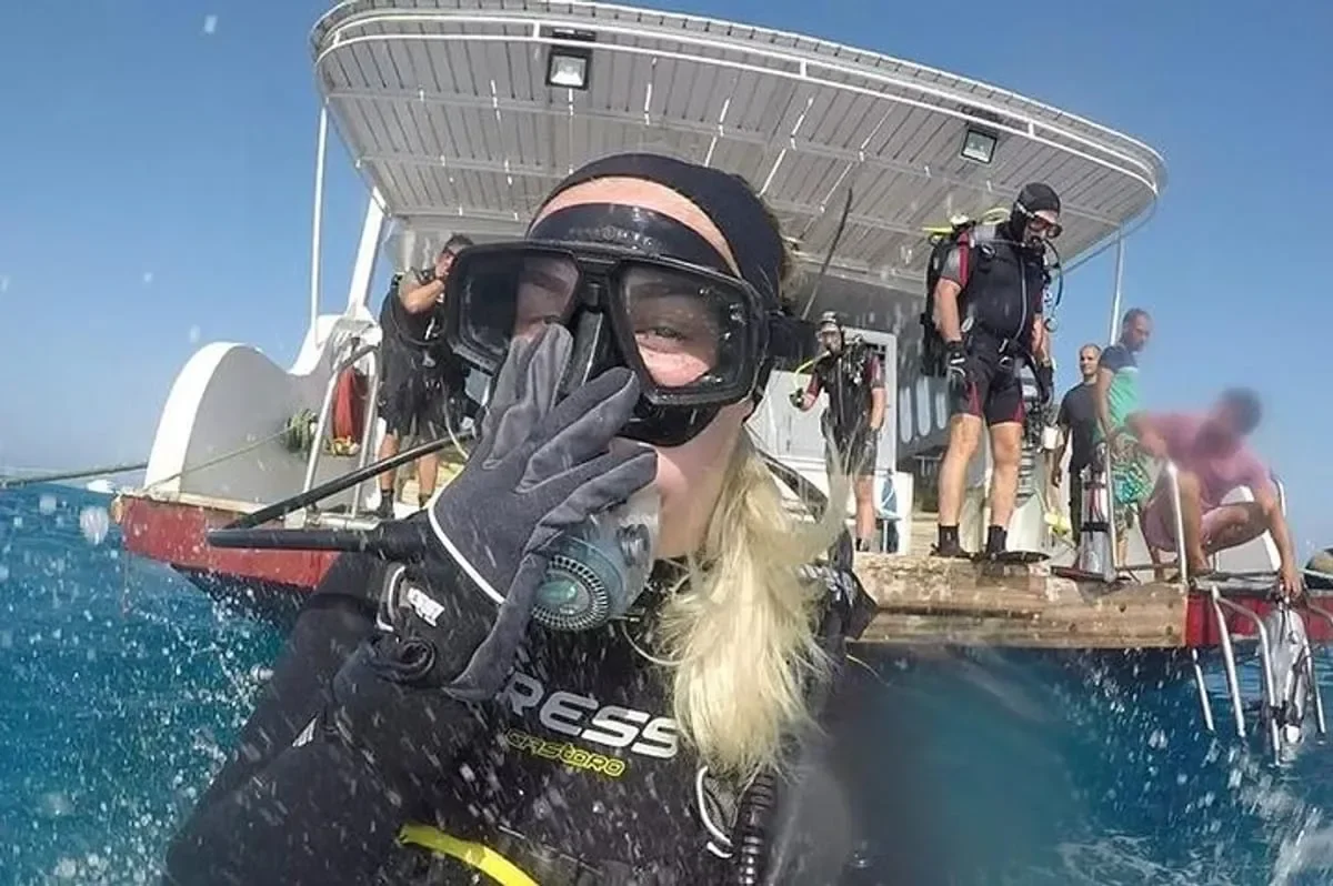 A diver wearing a mask and glove makes an OK hand sign close to the camera while other divers gear up on a dive boat's