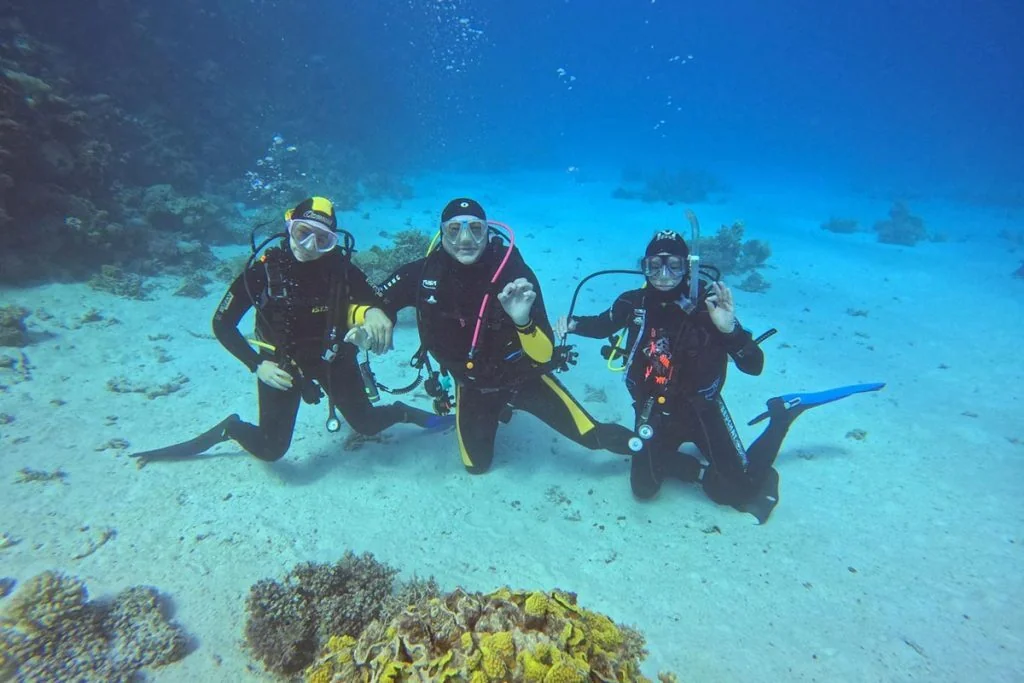 Three scuba divers kneel together on a sandy seafloor, each making an OK or peace hand sign toward the camera