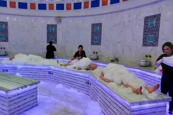 Several people wrapped in white foam and towels lie on a heated marble hammam platform in a domed tiled room