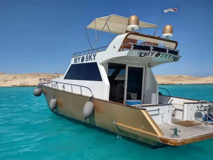 Close view of My Sky's bow and flybridge, with gold-topped fittings and a canvas sunshade, anchored off a sandy shore.
