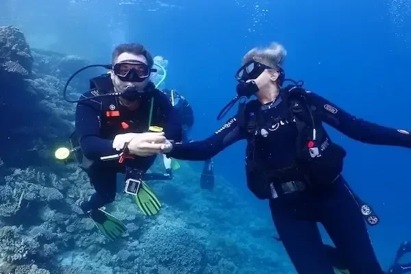 一名潜水教练和一名学员在岩石礁附近的水下面对面，互相打手势