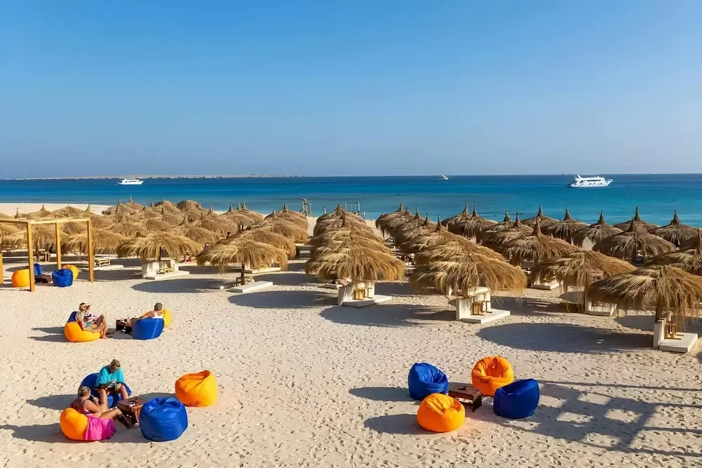 Strandclub met rijen rieten parasols en kleurige zitzakken op het zand, boten zichtbaar voor de kust
