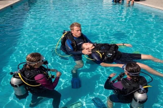 Ausbilder zeigt im Schwimmbecken den Kopfstützgriff an einem Schüler, zwei weitere Schüler in Tauchausrüstung sehen zu