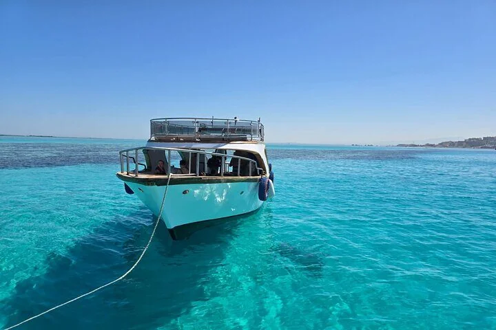 Bateau blanc à fond de verre ancré en eau turquoise peu profonde, pont supérieur vide, côte au loin
