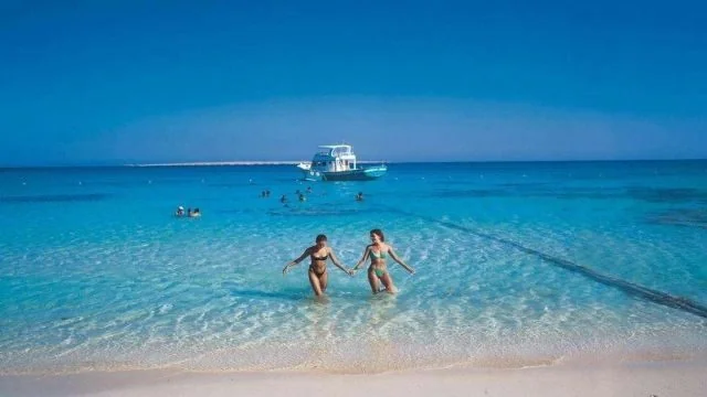 Két bikinis nő kéz a kézben sétál sekély türkizkék vízben a part felé, mögöttük hajóval és úszókkal