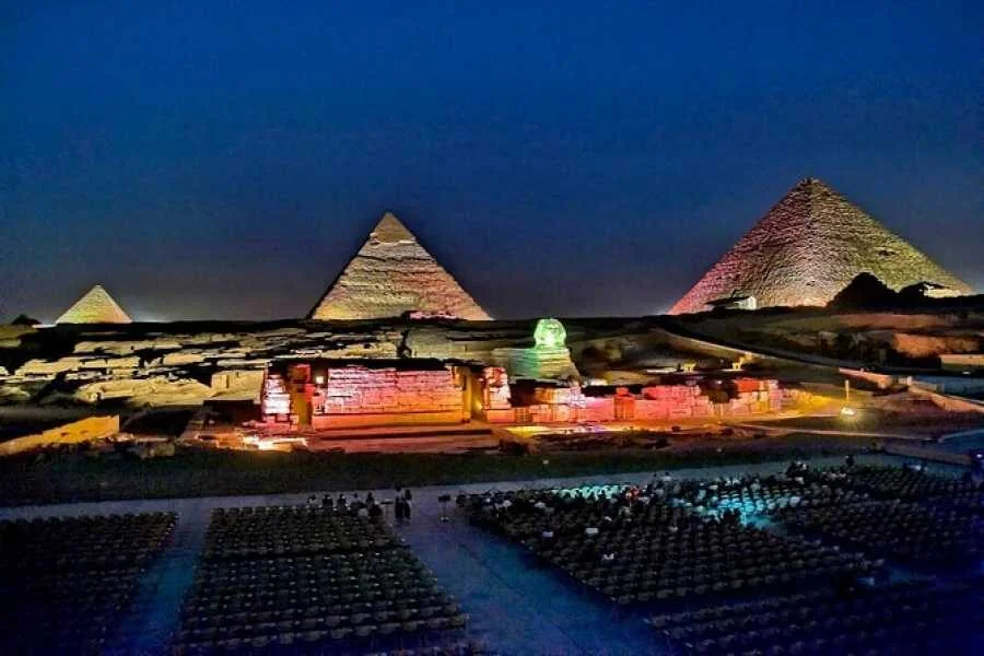 منظر ليلي لأهرامات الجيزة وأبو الهول مضاءةً، وأمامها صفوف من مقاعد الجمهور الفارغة