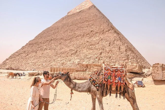 A couple pets a decorated camel with tasseled saddle blankets in front of the Great Pyramid of Khafre at Giza.