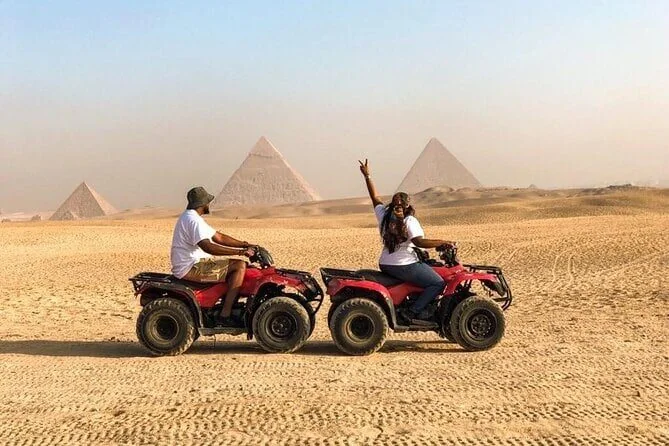 Twee rijders op rode quads door de open woestijn, één maakt een vredesteken, met twee piramides erachter