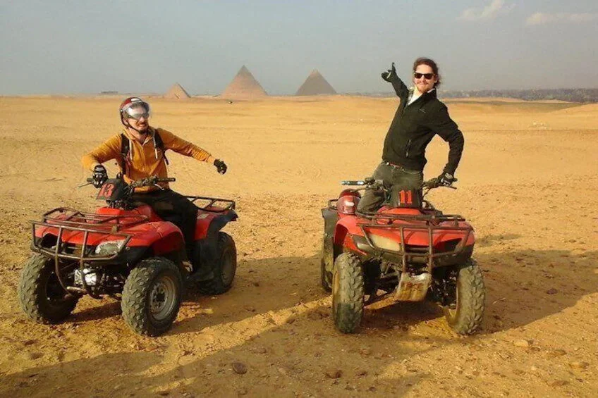 Twee mannen poseren op quads in de woestijn, één met duim omhoog, met twee piramides in de wazige verte