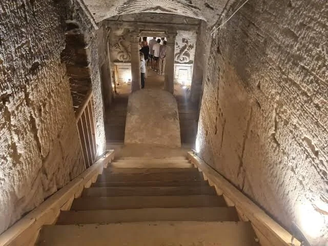 درج حجري يؤدي إلى أسفل في مقابر كوم الشقافة تحت الأرض، بجدران منحوتة وسياح ظاهرين في الأسفل