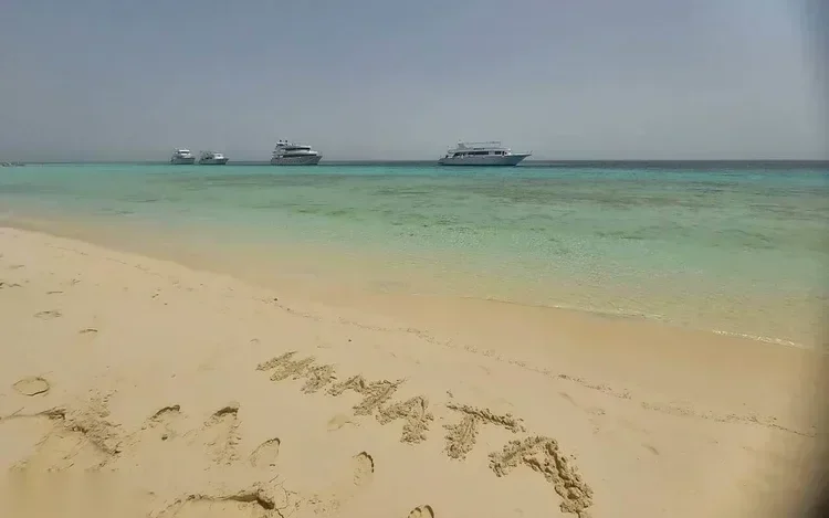 白色沙滩上用沙子写成的HAMATA字样，近海绿松石色海水中抛锚着几艘游览船