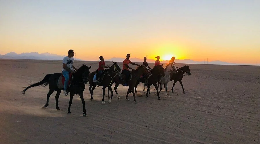 Silhouetted riders on horseback crossing a desert plain at sunset with mountains in the distance.