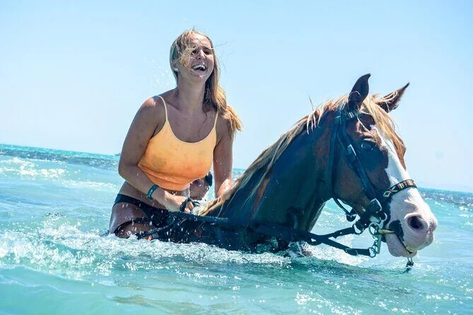 A woman laughing while standing chest-deep in the sea next to a brown-and-white horse.