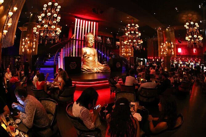 Schwach beleuchtete Lounge mit großer goldener Buddha-Statue, rotem Licht, Kronleuchtern und Gästen an Tischen