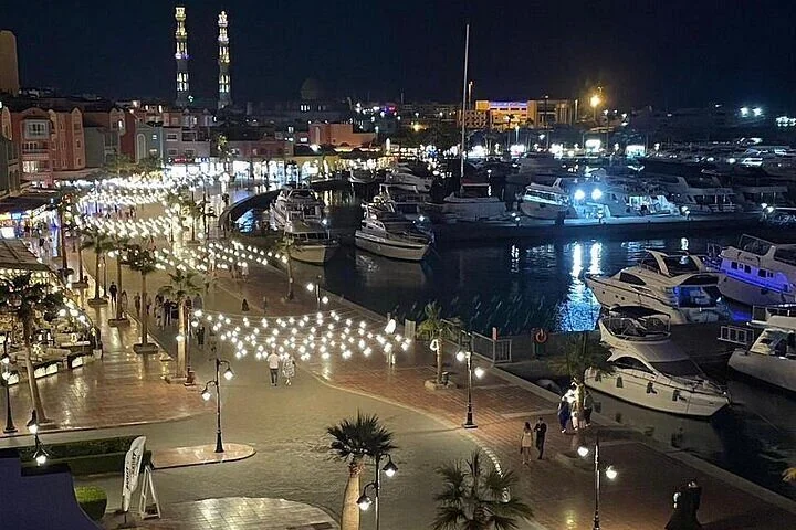 Nachtluftbild einer lichtergeschmückten Marina-Promenade mit festgemachten Booten, dahinter beleuchtete Minarette