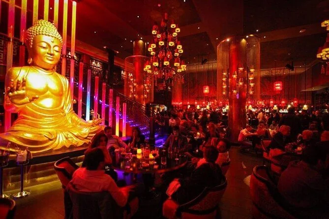 Volle Lounge-Bar bei Nacht mit großer goldener Buddha-Statue, roten Kronleuchtern und Gästen an Tischen