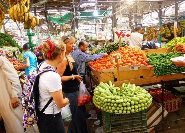 سياح يتجولون في سوق مغطى للفواكه والخضروات يعرض الموز والطماطم والخيار والفلفل