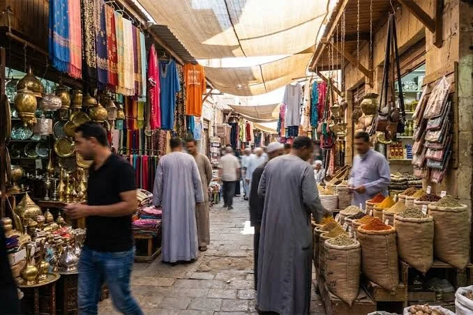 زقاق سوق مظلل تعلق فيه أدوات نحاسية ومنسوجات وأكياس توابل، وباعة ومتسوقون يمرون