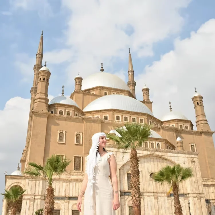 A woman in a white dress standing in front of the domed Mosque of Muhammad Ali, flanked by palm trees.