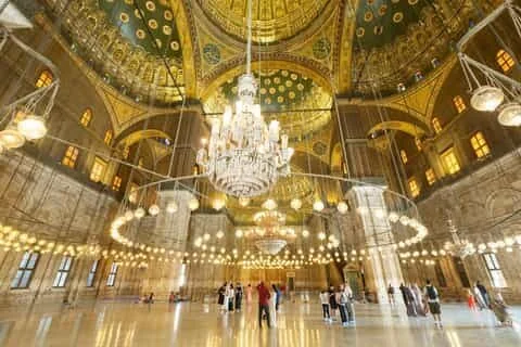 The ornately painted interior dome of a mosque with a large tiered chandelier and visitors below on the marble floor.