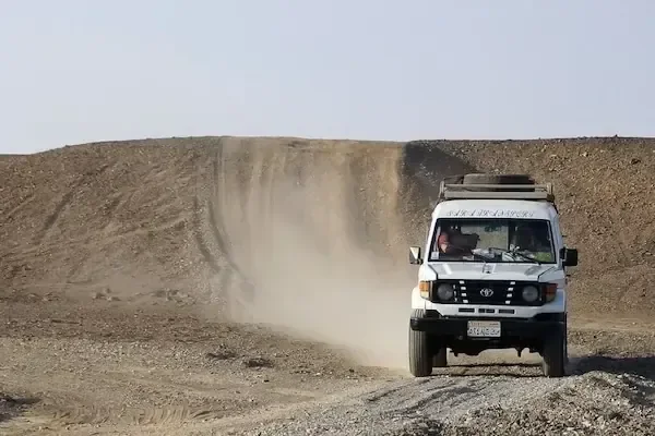 一辆载着两名乘客的白色四驱吉普车沿着尘土飞扬的沙漠小道行驶，身后扬起尘土
