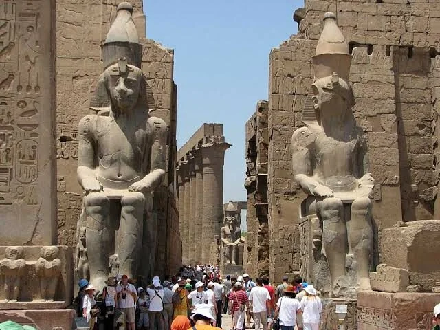 Two giant stone statues of a seated pharaoh wearing the double crown flank a crowded temple walkway at Karnak.