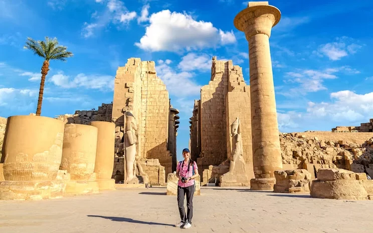A woman in a blue cap and backpack poses in front of ancient statues and columns at Karnak Temple under a blue sky.