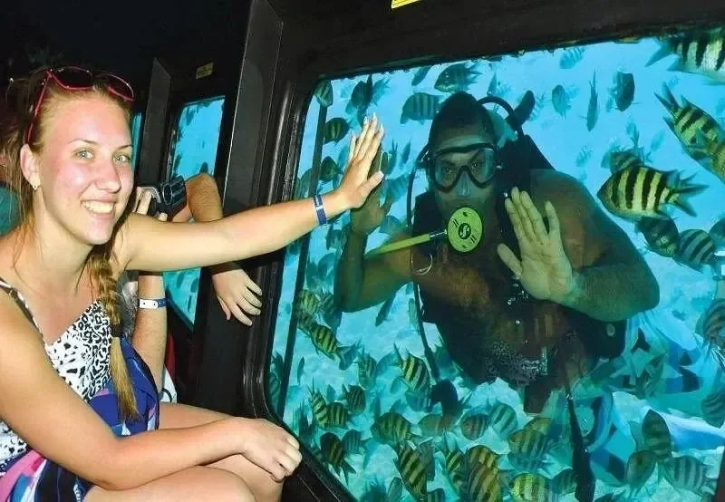 Mädchen gibt durch das Unterwasserfenster eines Halbtauchboots einem Taucher ein High Five, umgeben von gestreiften Fischen