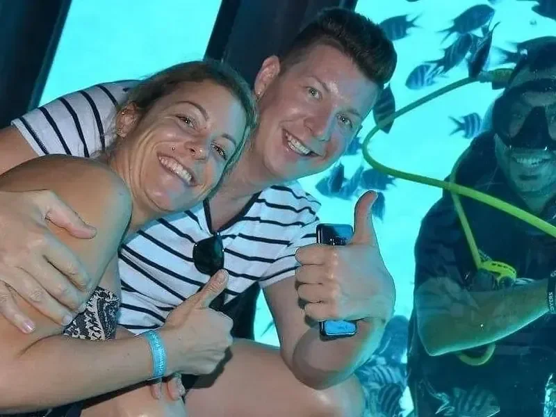 Zwei Freunde zeigen in der Kabine des Halbtauchboots Daumen hoch, hinter ihnen im Fenster ein Taucher und Fische